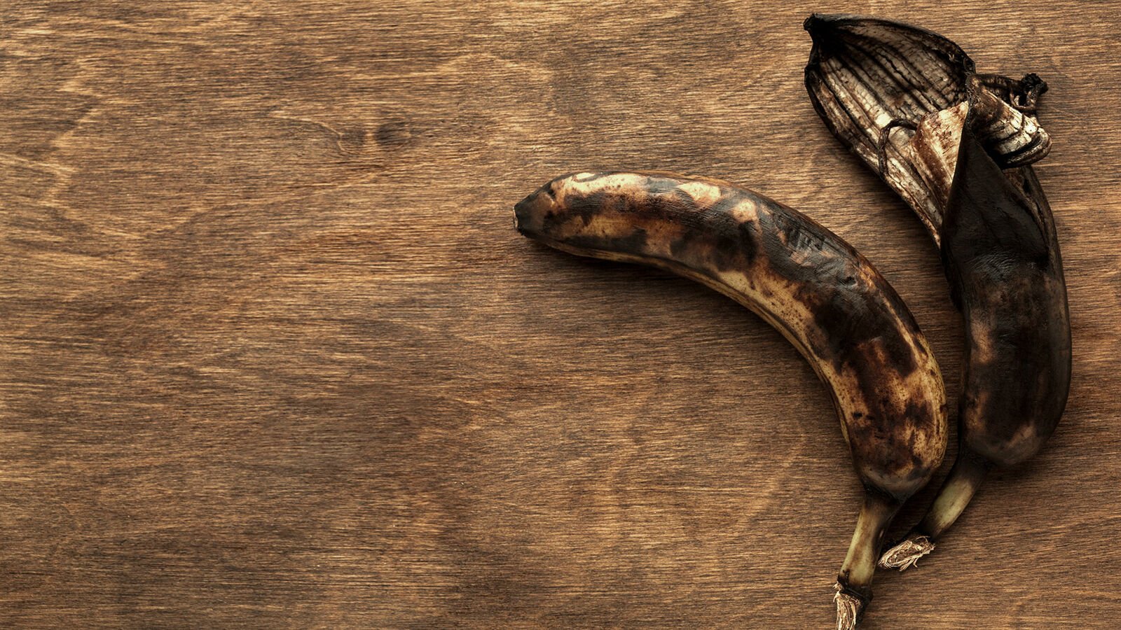 Two spoilt bananas with black peel lie on a wooden chopping board