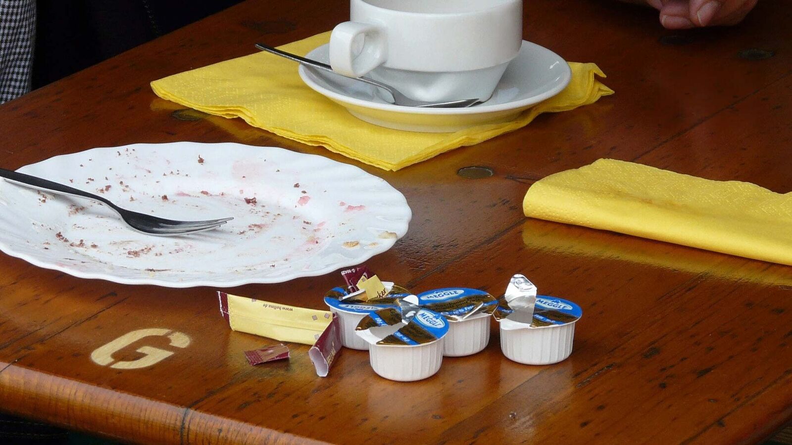 Prohibited: Mini portions with coffee milk in disposable plastic packaging On a table, next to a coffee cup, an empty cake plate and two yellow napkins, there are four open mini packets of coffee milk and a stick of sugar