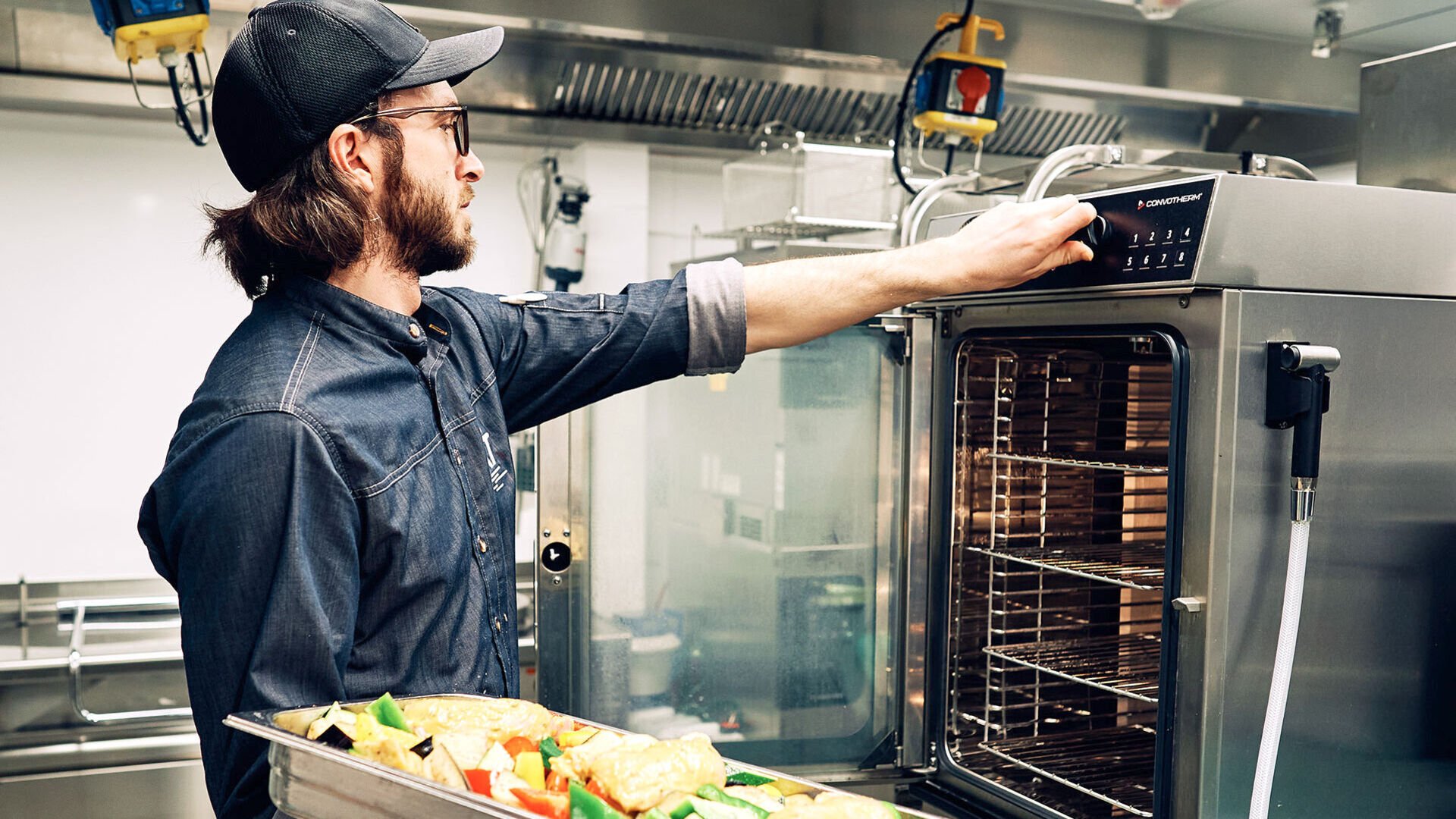 Energiespar-Tipp: Kombidämpfer Ein Koch steht mit einem Blech voller Gemüse am geöffneten Kombidämpfer und stellt die Temperatur ein