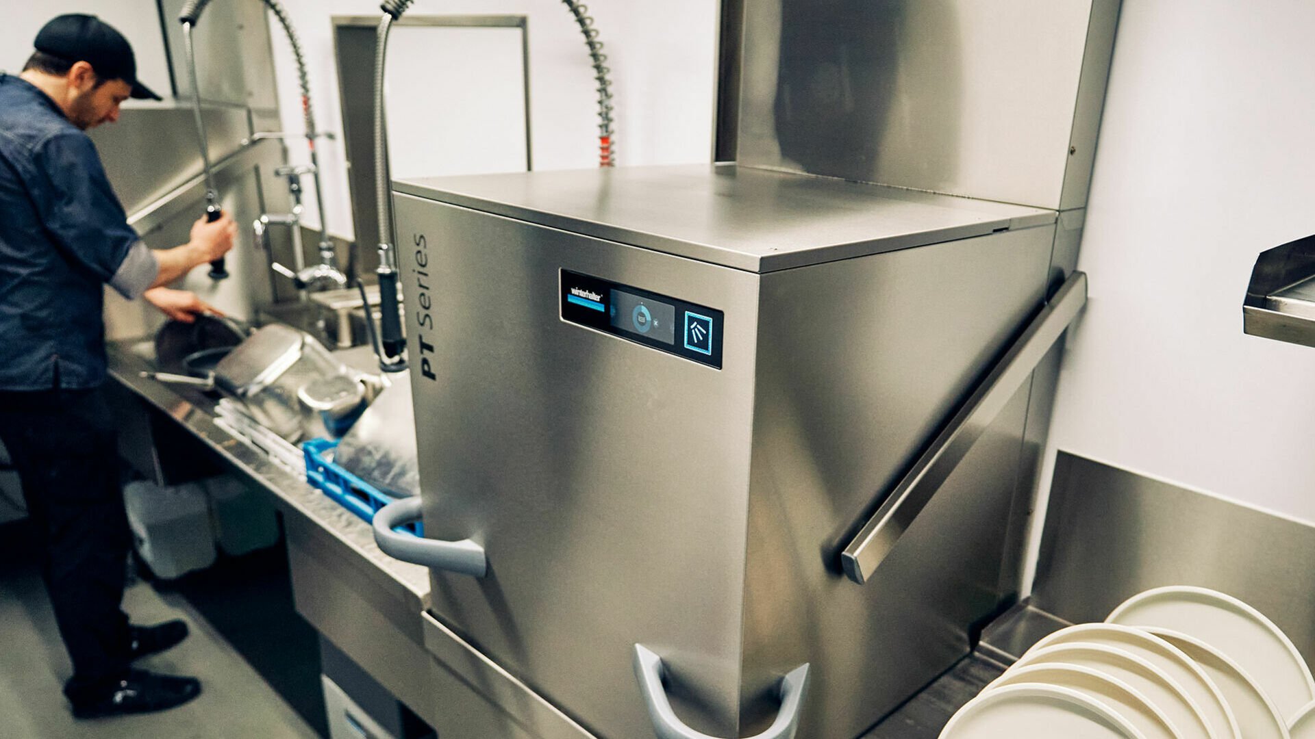 Energiespar-Tipps: Spültechnik In einer Spülküche läuft eine Haubenspülmaschine der PT-Serie von Winterhalter während im Hintergrund ein Mitarbeiter mit der Schlauchpendelbrause GN-Behälter vorspült