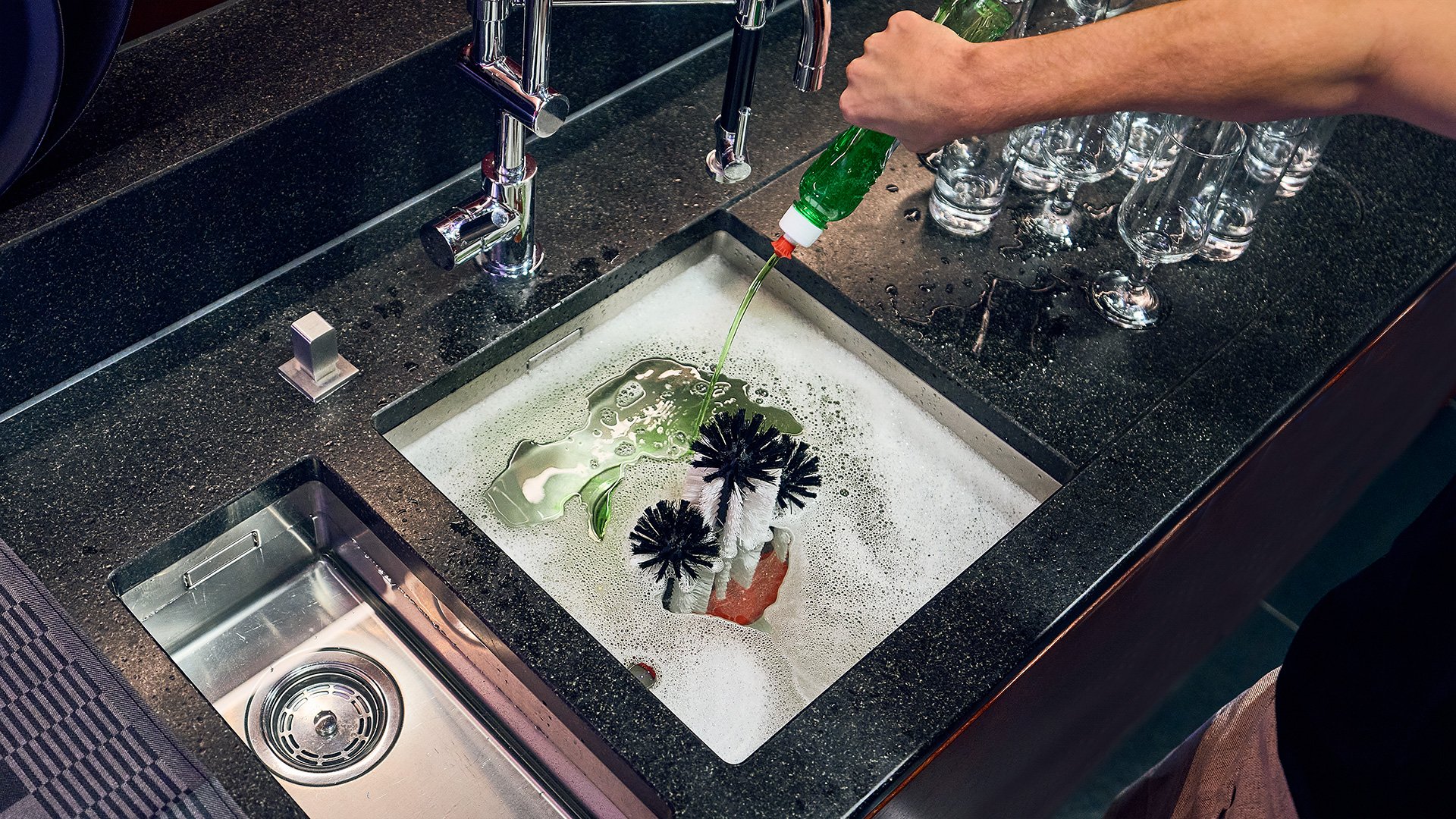 Inconvénient du lavage des verres à la main : dosage imprécis de lessive Un homme se tient devant l'évier d'un comptoir de bar et verse du détergent d'une bouteille dans l'eau