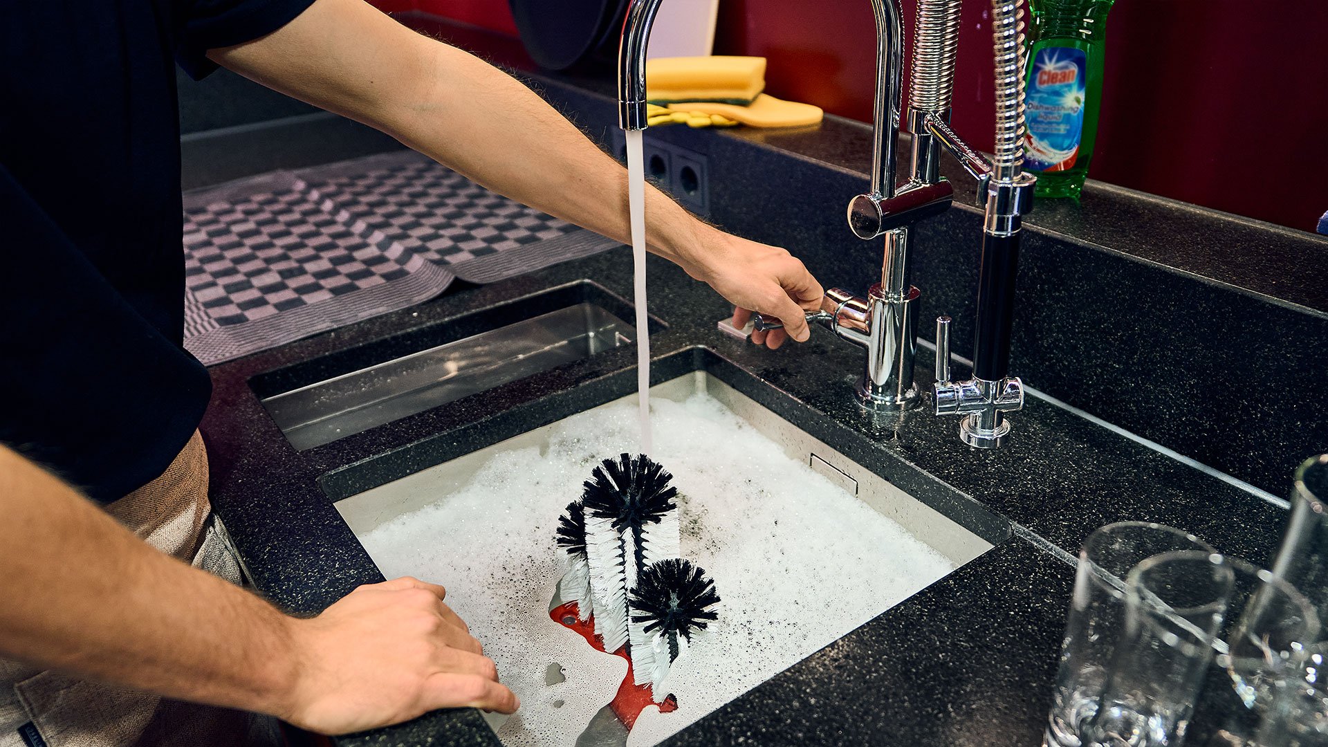 Inconvénient du lavage des verres à la main : forte consommation d'eau Un homme se tient derrière un comptoir de bar devant deux éviers remplis à ras bord d'eau et ferme le robinet
