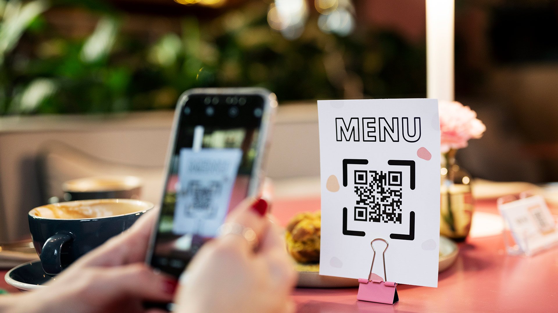 Digitale Speisekarte nutzen! Am Tisch eines Cafés hält eine Frau ihr Smartphone vor einen Aufsteller mit dem QR-Code zur digitalen Speisekarte