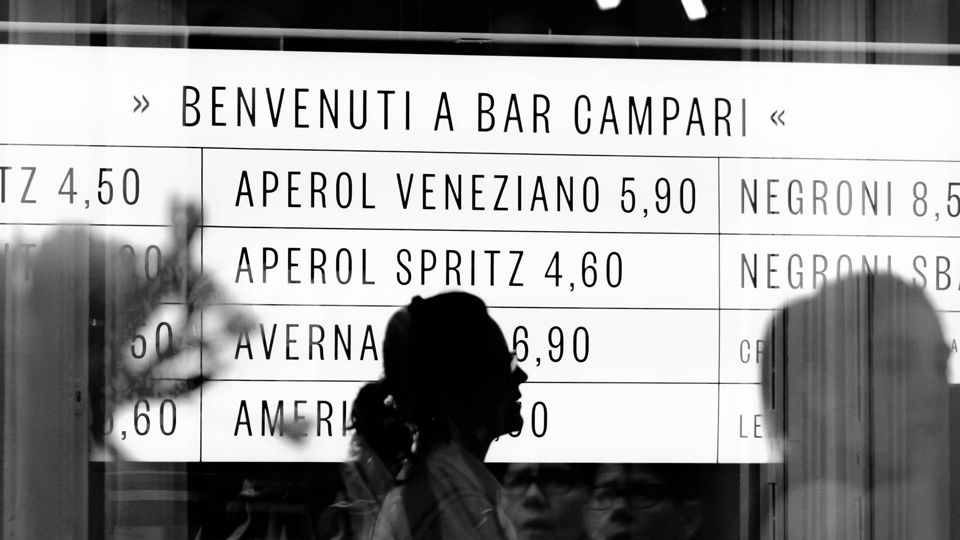 Das Angebot schmal und übersichtlich halten! Vor der hell erleuchteten Wand mit dem Getränkenagebot einer Bar sind unscharf einige Gäste zu erkennen