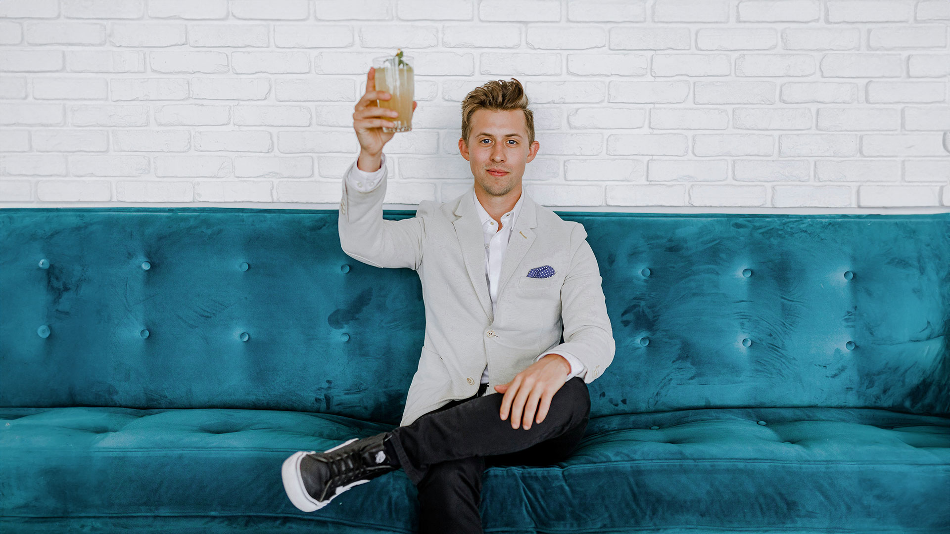 Mistake: Wrong ideas A young man in a blazer and sneakers sits on a sofa, smiling confidently and toasting the camera with a cocktail