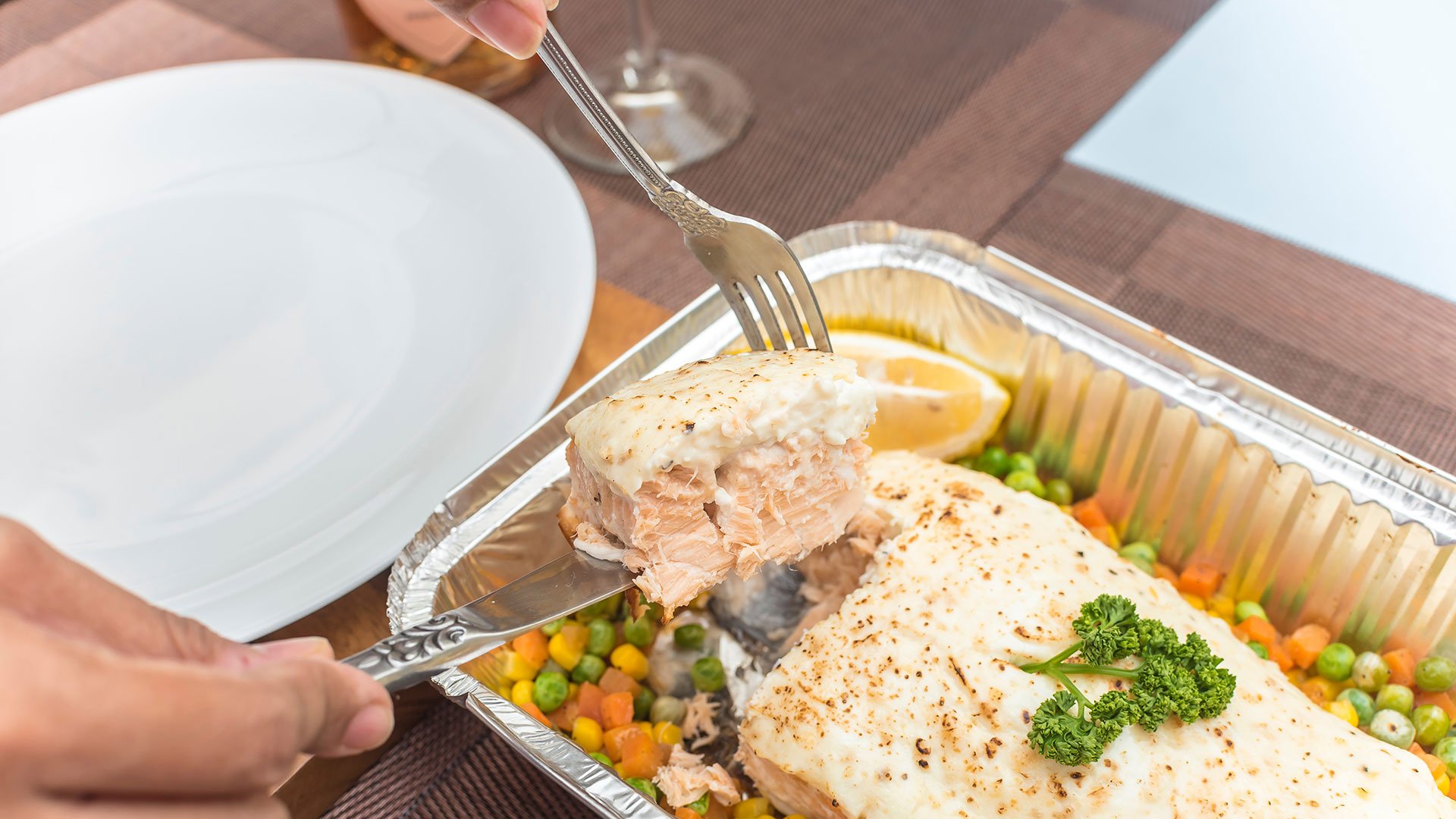 Mehrwegangebotspflicht für weitere Verpackungsmaterialien Aus einer Aluschale mit gebratenem Fischfilet und Gemüse wird mit Messer und Gabel ein Stück Fisch entnommen, um das Gericht auf einem weißen Teller anzurichten