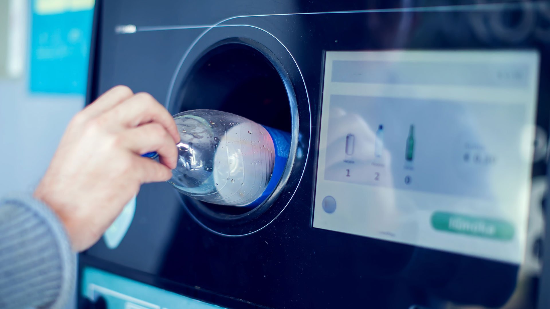 Rücknahmepflicht für Mehrweg-Getränkeverpackungen Eine Hand schiebt eine durchsichtige Mehrweg-Wasserflasche in die kreisrunde Öffnung eines Rückgabeautomaten für Pfandflaschen