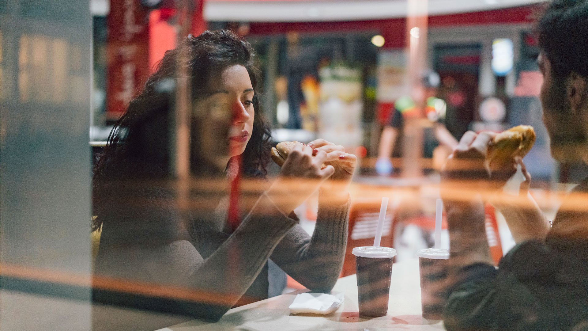 Verbot von Einwegverpackungen beim Vor-Ort-Verzehr Blick in ein Fastfood-Restaurant: Hinter der Scheibe sitzt ein junges Paar am Tisch und isst einen Burger